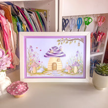 Load image into Gallery viewer, A framed iris fold picture card featuring a whimsical purple and cream toadstool house scene, displayed on a desk in a craft room setting with scissors, ribbons, and stationery in the background.