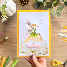Load image into Gallery viewer, A handmade greeting card featuring an iris fold fairy in a green dress sitting on a toadstool, held by a hand over a wooden desk with craft supplies.
