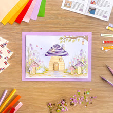 Load image into Gallery viewer, A handmade paper craft A4 card featuring an iris fold toadstool house, displayed on a wooden desk surrounded by craft supplies like coloured paper strips, sequins, and an instruction book.