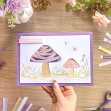 Load image into Gallery viewer, A handmade layered paper card featuring two iris fold toadstools in purple, pink, and cream, held by a hand over a wooden craft table surrounded by colourful paper strips, flowers, and scissors.