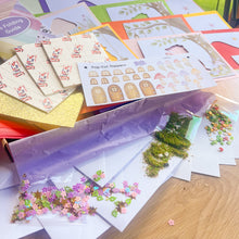 Load image into Gallery viewer, A craft kit spread out on a wooden table, including an iris folding guide, pop-cut toppers sheet with toadstool shapes, glitter card, moss, tissue paper, and small decorative sequins.
