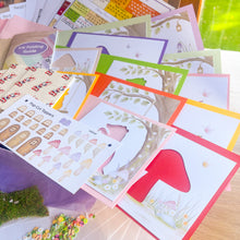 Load image into Gallery viewer, A collection of colourful handmade card-making crafting supplies, including a toadstool-themed card with a red toadstool cutout, card blanks and cut-outs, a pop-cut topper sheet, and an iris folding guide, arranged on a tabletop.