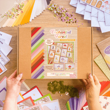 Load image into Gallery viewer, A person's hands hold a brown cardboard box labeled 'Enchanted Forest' craft kit, surrounded by colorful craft supplies, paper fans, flowers, and small decorative pieces on a wooden tabletop.
