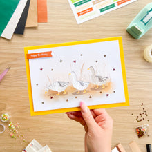 Load image into Gallery viewer, A hand holds a handmade greeting card featuring three white iris fold geese walking on a sandy path with small red leaves, set against a wooden desk with craft supplies.