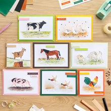 Load image into Gallery viewer, A flat-lay arrangement of eight handmade greeting cards featuring farm animals, displayed on a light wood background with crafting supplies like scissors, sequins, and paper around them.