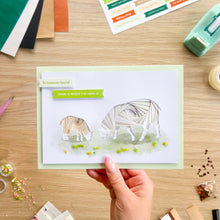 Load image into Gallery viewer, A handmade greeting card featuring an iris folding sheep and lamb on a white background with a light green border, held by a hand over a wooden craft desk surrounded by crafting supplies.