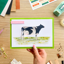 Load image into Gallery viewer, A hand holds a handmade greeting card featuring a black and white iris fold cow on a green background. The card has a pink banner that reads 'Have a brilliant Birthday!'.