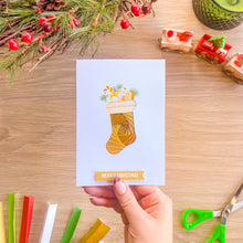 Load image into Gallery viewer, A hand holds a white Christmas greeting card featuring an iris fold golden Christmas stocking with wrapped gifts and candy canes, above a 'MERRY CHRISTMAS' banner. The card is photographed from above on a light wood table surrounded by festive props including pine branches, red berries, a toy train, and green-handled scissors.