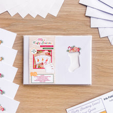 Load image into Gallery viewer, A packaged craft kit labeled 'Mini Fluffy Stocking Card Aperture Pack Embellished Edition' sits on a light wood surface, surrounded by white paper envelopes and aperture cards. The package shows a white stocking-shaped cut-out and colorful front insert graphics.