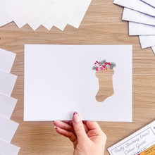 Load image into Gallery viewer, A hand holds a white greeting card with a Christmas stocking cut-out, filled with small illustrated gifts and evergreen branches, on a light wood tabletop. More white cards are fanned out around it.