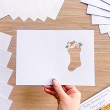 Load image into Gallery viewer, A hand holds a white greeting card with a Christmas stocking cut-out, filled with small illustrated gifts and evergreen branches, on a light wood tabletop. More white cards are fanned out around it.