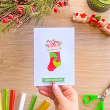 Load image into Gallery viewer, A hand holds a white Christmas greeting card featuring a colorful iris fold Christmas stocking with gifts and the text 'SANTA'S ON HIS WAY!'. The card is photographed from above on a light wood tabletop with holiday decorations, paper strips, and small toy trains around it.