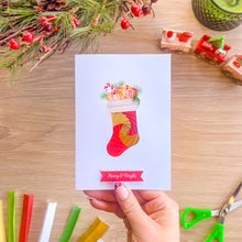 Load image into Gallery viewer, A hand holds a white Christmas greeting card featuring a colourful iris fold stocking filled with presents and pine branches, with a red 'Merry & Bright' banner at the bottom. The card is photographed on a wooden tabletop with holiday decorations, scissors, and craft strips around it.