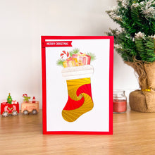 Load image into Gallery viewer, Christmas card with a red border featuring a yellow stocking filled with gifts, placed on a wooden surface with festive decorations.