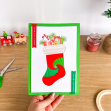 Load image into Gallery viewer, Hand holding a Christmas card with a red and green stocking on a wooden surface.