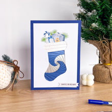 Load image into Gallery viewer, Framed Christmas card with blue stocking and presents on a wooden surface with decorative items.