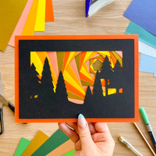 Load image into Gallery viewer, A hand holds a square layered paper art card featuring black tree silhouettes against a spiral background of orange, yellow, and pink paper. The card has a bright orange border and is photographed on a wooden desk with craft supplies around it.