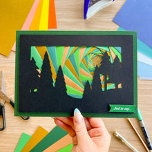 Load image into Gallery viewer, A handmade layered card featuring black silhouette trees against a vibrant green, orange, and teal spiral background, held by a hand over a wooden craft table with colorful paper sheets.