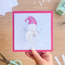 Load image into Gallery viewer, A handmade square greeting card with a bright pink border and a white snowman design made by iris folding paper, held by a hand over a wooden table with crafting tools.