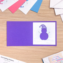 Load image into Gallery viewer, An open purple card blank on a light wood surface, showing a white aperture with a purple snowman illustration wearing a Santa hat. Partially visible pattern sheets and envelopes surround it, with a package labeled 'Frosty Snowman' in the lower right.