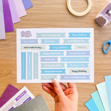 Load image into Gallery viewer, Sheet of printed phrases for cards held by a hand on a wooden surface with card-making supplies.
