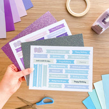 Load image into Gallery viewer, Sheets of glitter card and pre-printed sentiments held by a hand on a wooden surface with stationery items.