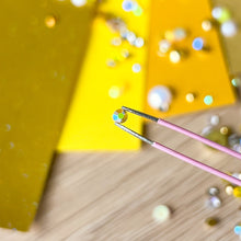 Load image into Gallery viewer, Close-up of tweezers with a small gem on a wooden surface with yellow paper and more gems in the background.