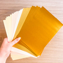 Load image into Gallery viewer, A hand holds a fanned stack of metallic gold and cream-colored sheets of craft paper, on a light wood grain background.