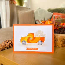 Load image into Gallery viewer, A greeting card with an orange iris fold truck carrying hay bales, displayed on a wooden table in a cozy autumn-themed setting with pinecones and fall decor.