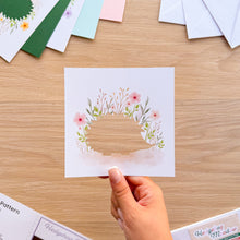 Load image into Gallery viewer, A hand holds a square white card featuring watercolor flowers around a cutout hedgehog shape, photographed over a light wood tabletop with other cards and envelopes scattered around.