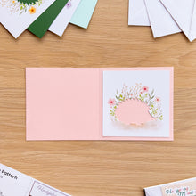Load image into Gallery viewer, A pink card blank with a hedgehog aperture card with printed flower design in pink, green, and peach tones.