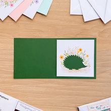 Load image into Gallery viewer, A dark green card blank with a hedgehog aperture card with yellow flowers, displayed on a light wood tabletop.