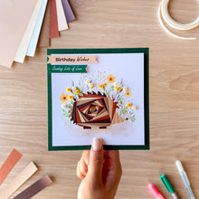 Load image into Gallery viewer, A handmade square greeting card held by a hand on a light wood desk. The card features a dark green border, a white center, and an iris fold hedgehog made of layered brown paper, surrounded by yellow and white wildflowers.