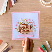 Load image into Gallery viewer, An iris fold hedgehog on a pink card blank embellished with pink and white flowers, held by a hand over a wooden desk with craft supplies.