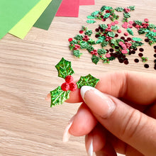 Load image into Gallery viewer, A hand with manicured nails holds a small, shiny green fabric holly leaf ornament with red berries, set against a light wood background scattered with more decorative holly pieces and colored paper sheets.