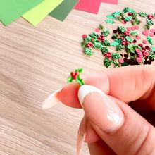 Load image into Gallery viewer, A close-up of a hand with manicured nails holding a tiny green and red resin holly ornament, with more of the same decorative pieces scattered on a light wood surface in the background.