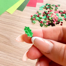 Load image into Gallery viewer, A hand with manicured nails holds a small, shiny green holly leaf foil embellishment. In the background, scattered red and green Christmas glitter decorations and colorful paper sheets are visible on a light wood surface.