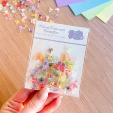 Load image into Gallery viewer, Packaged colourful butterfly embellishments held by a hand on a wooden surface with additional embellishments scattered around.