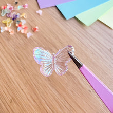 Load image into Gallery viewer, Butterfly-shaped object on a wooden surface with pink tweezers.