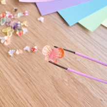 Load image into Gallery viewer, Butterfly-shaped object on a wooden surface with pink tweezers.