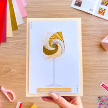 Load image into Gallery viewer, Greeting card with wine glass design held by a hand on a wooden table with craft supplies.
