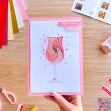 Load image into Gallery viewer, Hand holding a birthday card with a pink cocktail design on a wooden table.