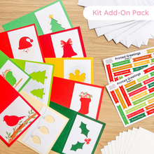 Load image into Gallery viewer, A flat lay of a Christmas card kit add-on pack showing various colorful greeting cards with holiday illustrations like Santa Claus, a Christmas tree, a bell, a bird, holly, and a stocking, alongside sheets of printed holiday labels on a light wood surface.