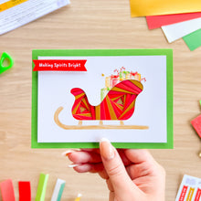 Load image into Gallery viewer, A hand holds a handmade Christmas greeting card featuring a red and gold patterned sleigh carrying gifts, with the text 'Making Spirits Bright' on a red banner. The card has a green border and is photographed on a wooden table with craft supplies scattered around.