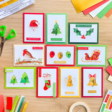 Load image into Gallery viewer, A flat-lay of ten Christmas greeting cards arranged on a light wood tabletop, surrounded by craft supplies including scissors, colored paper, tape, and markers. The cards feature colorful holiday illustrations such as a red Santa hat, green Christmas trees, holly, ornaments, a sleigh, and a golden angel.