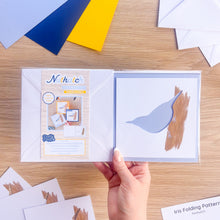 Load image into Gallery viewer, A hand holds a Nuthatch Aperture Pack card set on a light wood desk, showing a nuthatch bird-shaped card. Colorful paper sheets are scattered around the workspace.