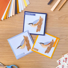 Load image into Gallery viewer, Three handmade iris fold nuthatch greeting cards arranged on a light wood tabletop with craft paper strips, scissors, and sticker sheets nearby.