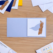 Load image into Gallery viewer, A Nuthatch aperture on a light blue greeting card blank, on a light wood tabletop. Partially visible cards, envelopes, and printed materials are arranged around it on the table.