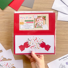 Load image into Gallery viewer, A hand holds a packaged 'Party Cracker' card aperture pack set on a wooden tabletop, surrounded by loose cards, envelopes, and craft papers. The package is red with a clear plastic sleeve and a white insert showing a red party cracker design with confetti.