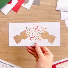 Load image into Gallery viewer, A hand holds a white paper cut-out of two cracker halves and colourful confetti, on a wooden tabletop with craft papers and envelopes around it.